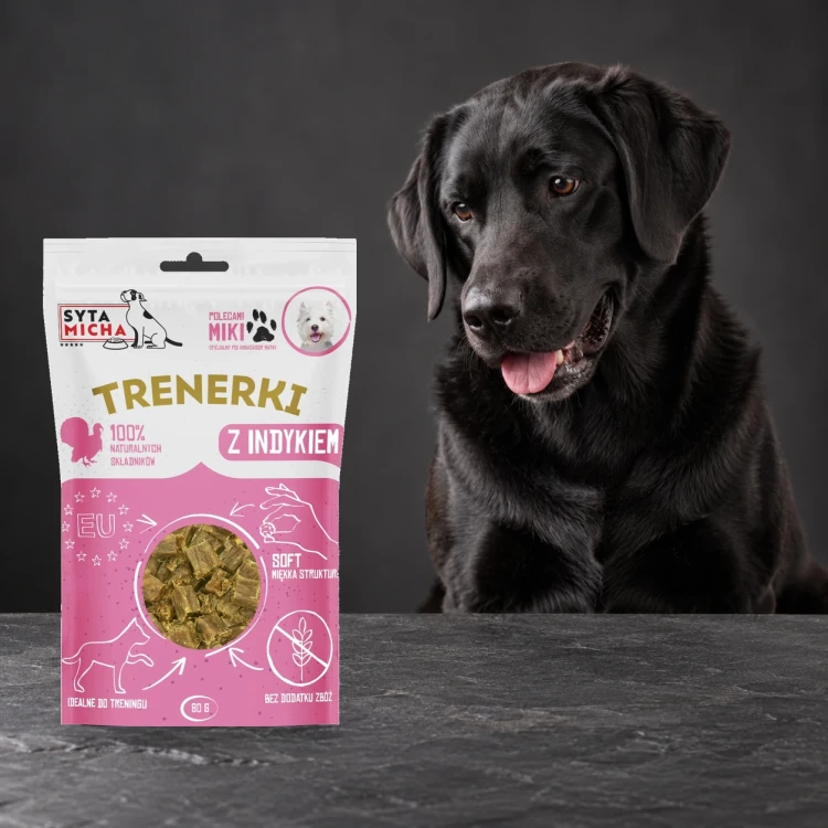 Trenerki Syta Micha z indykiem 80 g obok psa na ciemnym tle.