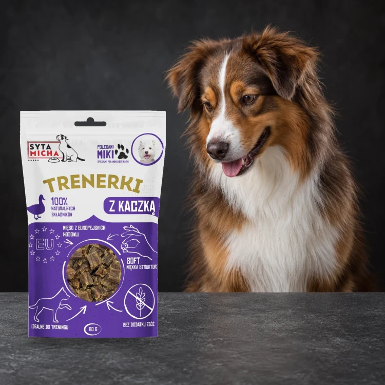 Trenerki Syta Micha z kaczką 80 g obok psa na ciemnym tle.
