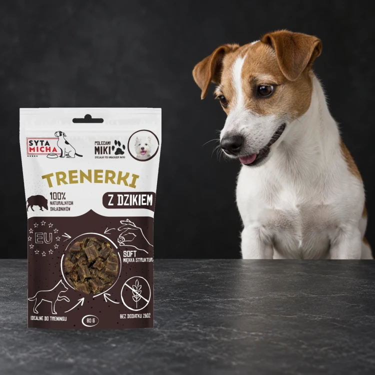 Trenerki Syta Micha z dzikiem 80 g obok psa na ciemnym tle.