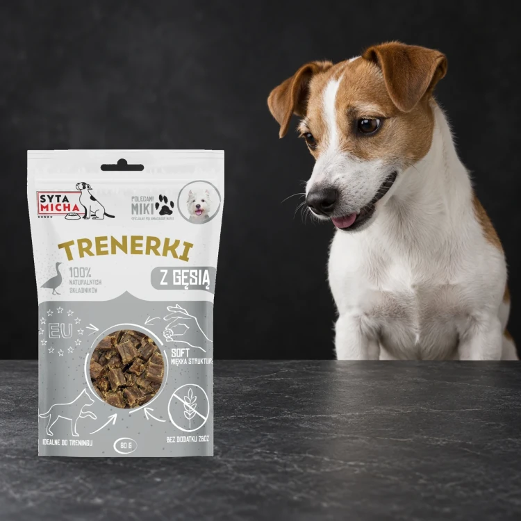 Trenerki Syta Micha z gęsią 80 g obok psa na ciemnym tle.
