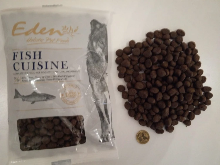 Mała saszetka Eden Fish Cuisine obok rozsypanych granulek oraz monety pokazującej skalę